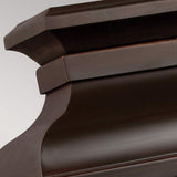 A close-up of dark wooden crown molding with an intricate design highlights the craftsmanship, featuring smooth curves and sharp edges with a polished, rich finish. This elegance is enhanced by the Pediment 2 Light Outdoor Medium Wall Lantern in Dark Copper, which adds a warm glow. The plain background accentuates the artisan quality of the woodwork.