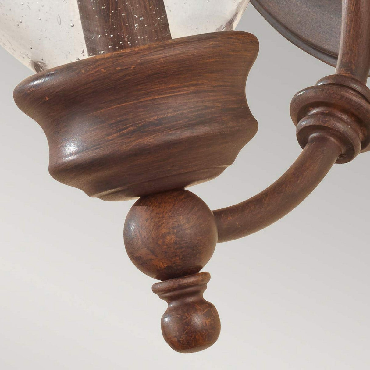 A close-up of the Pickering Lane 1 Light Wall Light - Bronze, featuring an antique-style design with a curved arm and glass shade. Emphasizing farmhouse aesthetics, this fixture includes intricate details such as a round knob on the arm. The background is softly blurred in light gray.