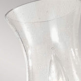 The detailed view of the Pickering Lane 1 Light Wall Light in bronze showcases its clear, textured glass design with a wide opening, reminiscent of a storm glass lantern. The presence of small bubbles within the glass enhances its charm, while the soft gray background provides contrast, making this fixture an elegant addition to any farmhouse décor.