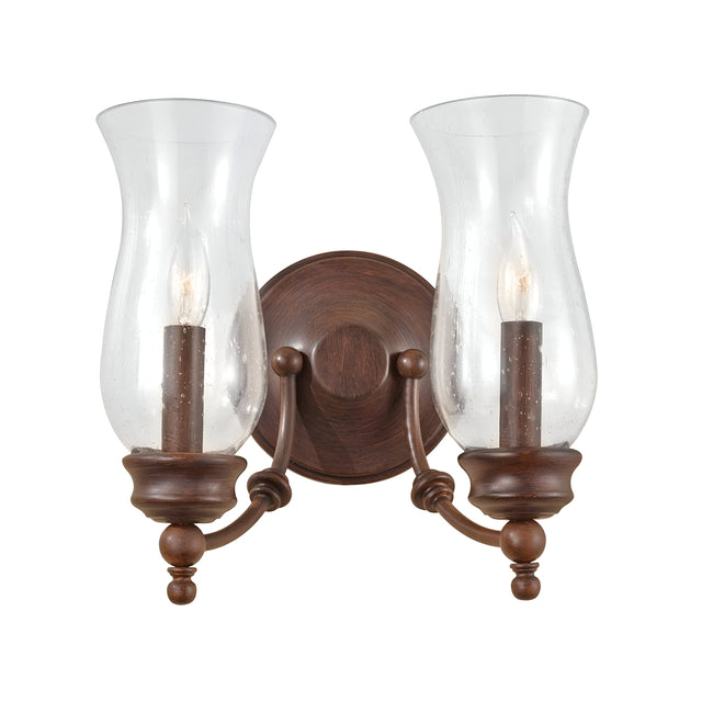 The Pickering Lane 2 Light Wall Light - Bronze is a vintage-style wall sconce with a traditional aesthetic. It features two glass hurricane shades and candle-shaped bulbs. This fixture, finished in Heritage Bronze, showcases curved arms and decorative finials reminiscent of storm glass lanterns, all attached to a circular backplate.