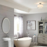 A modern bathroom features a freestanding oval bathtub, pedestal sink, and mirrored cabinet. The space, adorned in light gray tones with abstract art and sheer curtains, is elegantly illuminated by the Prospect Park 2 Light Semi Flush in Satin Nickel with Chrome. An opal etched glass mirror adds a chic touch.