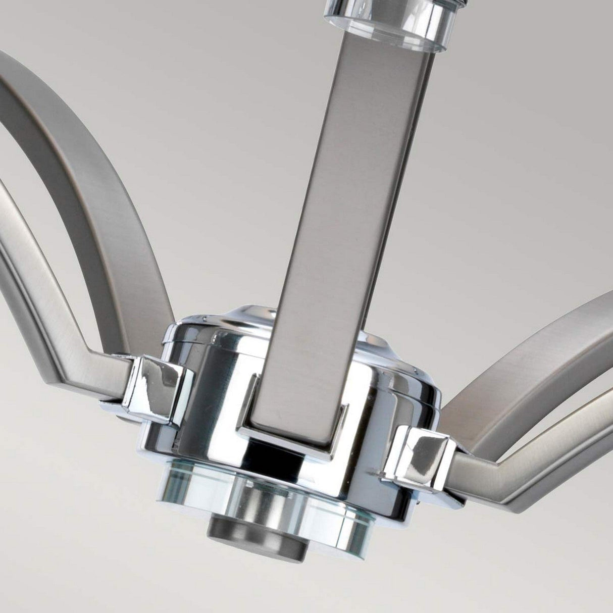 A close-up of the Prospect Park 5 Light Chandelier - Satin Nickel With Chrome highlights its modern elegance with sleek, curved arms and a polished finish. The central fixture and intersecting elements enhance its contemporary charm, making it ideal for transitional spaces against a light gray background.