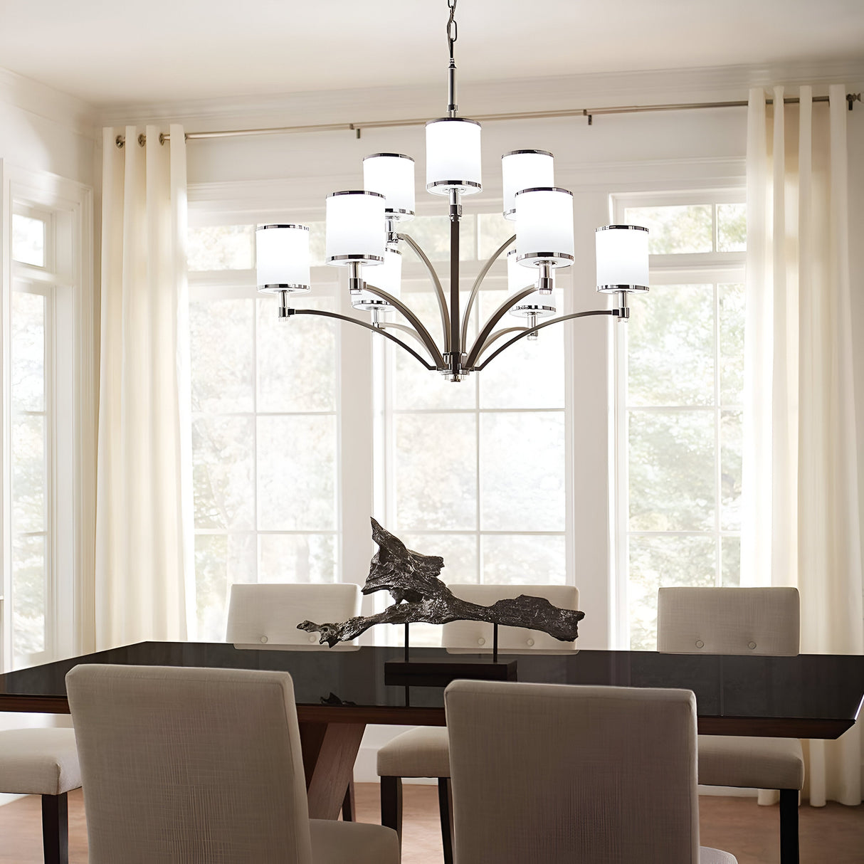 A modern dining room features a large window with sheer curtains, a black rectangular table, beige upholstered chairs, and a stunning Prospect Park 9 Light Chandelier in satin nickel with chrome, complete with opal etched glass lampshades illuminating an abstract sculpture centerpiece.