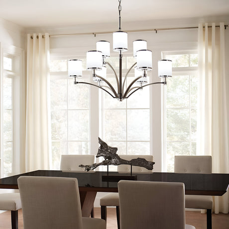 A modern dining room features a large window with sheer curtains, a black rectangular table, beige upholstered chairs, and a stunning Prospect Park 9 Light Chandelier in satin nickel with chrome, complete with opal etched glass lampshades illuminating an abstract sculpture centerpiece.