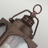 A close-up of the Randhurst 1 Light Outdoor Small Wall Lantern - Copper highlights its rustic, weathered copper oxide finish. The lantern boasts clear glass panels and decorative perforations near the top, accompanied by a loop handle for easy hanging. A soft gradient background enhances its vintage charm.