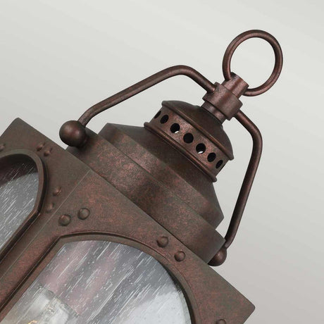 A close-up of the Randhurst 1 Light Outdoor Small Wall Lantern - Copper highlights its rustic, weathered copper oxide finish. The lantern boasts clear glass panels and decorative perforations near the top, accompanied by a loop handle for easy hanging. A soft gradient background enhances its vintage charm.