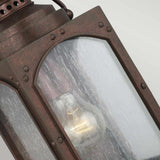 A close-up view of the Randhurst 1 Light Outdoor Small Wall Lantern showcases its antique flair with a visible light bulb inside. The lantern boasts a rectangular, weathered bronze frame and slightly textured glass panels. When illuminated, it emits a warm glow against its rugged composite construction, accentuated by the Copper Oxide finish.