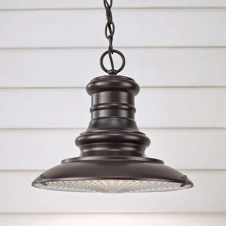 A Redding Station 1 Light Outdoor Medium Chain Lantern in dark bronze, with a vintage design, is suspended from a chain against a white paneled wall. This lamp includes a wide, round shade and features a textured glass diffuser at the bottom, echoing the classic charm of the Redding Station Light.