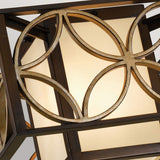 A close-up of the Remy 2 Light Flush in a Heritage Bronze finish with Parissiene Gold detailing highlights its geometric metal patterns layered over luminous frosted glass panels. The design casts a warm, ambient glow and presents an intricate circular motif that subtly echoes the elegance of French Arts and Crafts style.