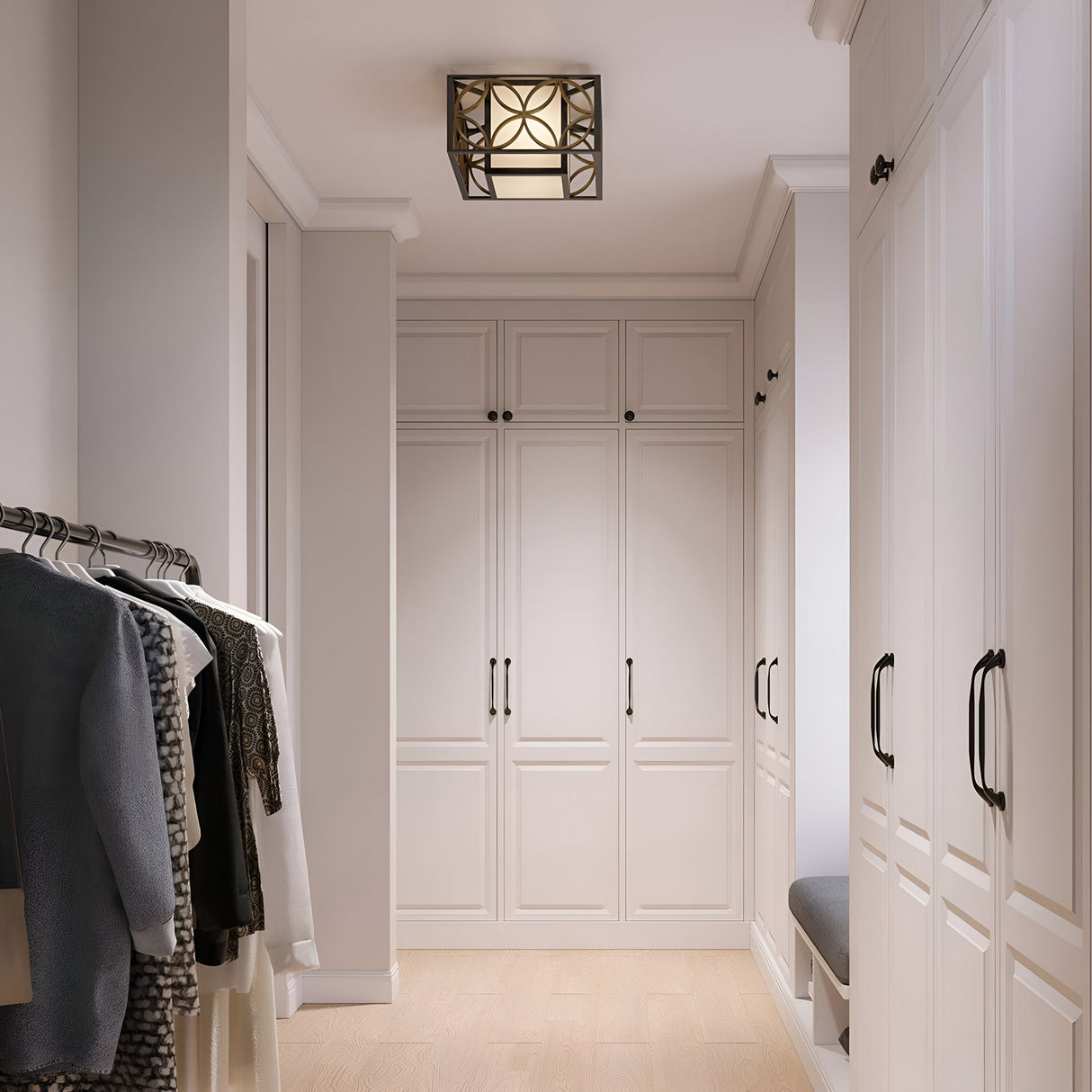 A walk-in closet with white cabinets and wooden flooring showcases clothes hanging on a rail to the left and a cushioned bench built into the cabinetry on the right. The Remy 2 Light Flush, designed in French Arts and Crafts style, emits an elegant glow from above with its Heritage Bronze/Parissiene Gold finish.