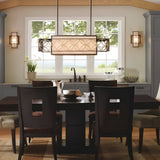 A modern dining room is highlighted by a dark wooden table surrounded by matching chairs, with the Remy 4 Light Pendant Chandelier in Heritage Bronze/Parissiene Gold serving as the central decorative fixture. A large window offers a scenic nature view, complemented by a vase of flowers, elegant glassware, and cabinetry in neutral tones.