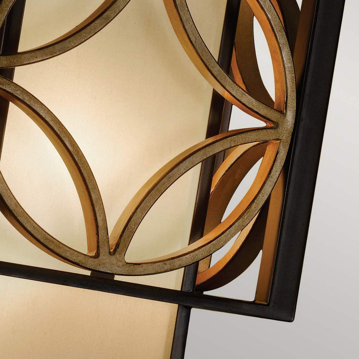 A close-up view of the Remy 1 Light Wall Light in Heritage Bronze/Parisienne Gold reveals a geometric design with overlapping circular patterns. Its metallic structure, combined with a warm, glowing backdrop, exudes an artistic and elegant appearance reminiscent of the French Arts and Crafts style.