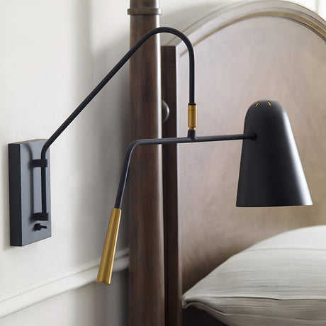 The Simon 1 Light Wall Light - Black, featuring an adjustable arm and a sleek black finish, is gracefully installed beside a wooden bedframe. Its gold accents enhance the sophisticated look as it extends outward, illuminating the neatly arranged bed with crisp white linens.