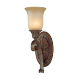 The Stirling-1 Castle 1 Light Wall Light - Bronze features a traditional wall sconce design with an intricate metal base and floral embellishments. Its upward-facing frosted glass shade emits a gentle, warm illumination, complemented by an antique bronze finish that brings rustic elegance to any setting.