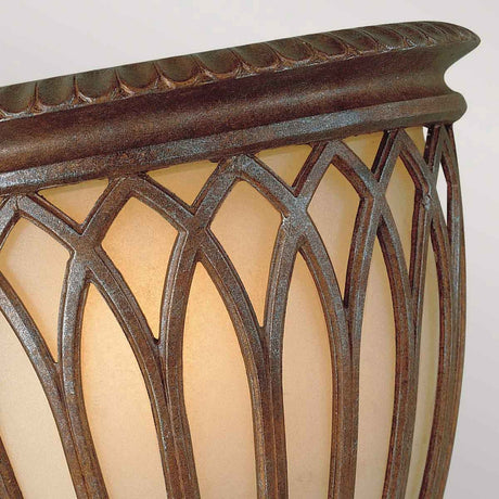 A close-up of the Stirling-2 Castle 1 Light Wall Light - Bronze reveals a decorative lamp with an intricate lattice design on its textured metal frame, completed with a bronze finish. The translucent shade casts a warm, soft light, enhancing the cozy ambiance reminiscent of traditional lighting, along with an ornate scalloped edge at the top of the frame.