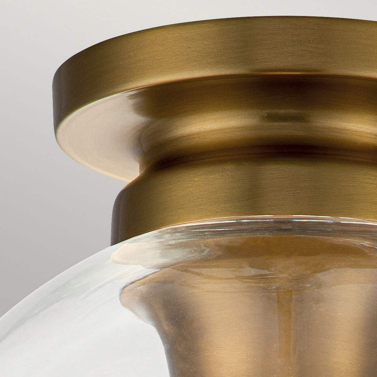 Close-up of a ceiling light fixture with a burnished brass finish. The Tabby 1 Light Flush - Burnished Brass showcases a clear glass dome and exposed lamping, connected to a rounded base, set against a neutral background.