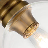 A close-up view of the Tabby 1 Light Flush in Burnished Brass showcases a vintage-style light bulb encased in a clear glass fixture. The illuminated bulb casts a warm glow, highlighting the antique finish of the metal socket. This design elegantly features an exposed lamping aesthetic for an exquisite touch.