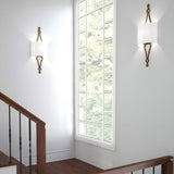 A bright stairwell with wooden steps and a black railing features two Tilling 1 Light Wall Lights in Antique Gold, each showcasing a white linen shade. Mounted on a white wall, these sconces complement the large window that offers a view of the lush green foliage outside.