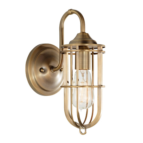 The Urban Renewal 1 Light Wall Light in Dark Antique Brass showcases a vintage-style design with an exposed Edison-style bulb encased in a metal cage. It features a round backplate and an upward-curving arm, combining industrial elegance with antique brass for a timeless appearance.