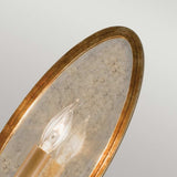 A close-up of the Valentina 1 Light Wall Light - Oxidized Bronze highlights its decorative fixture with two exposed candle-shaped bulbs. The background reveals an oval frame with a golden metallic edge and a textured surface, adorned with polished glass crystals to create an elegant vintage aesthetic.