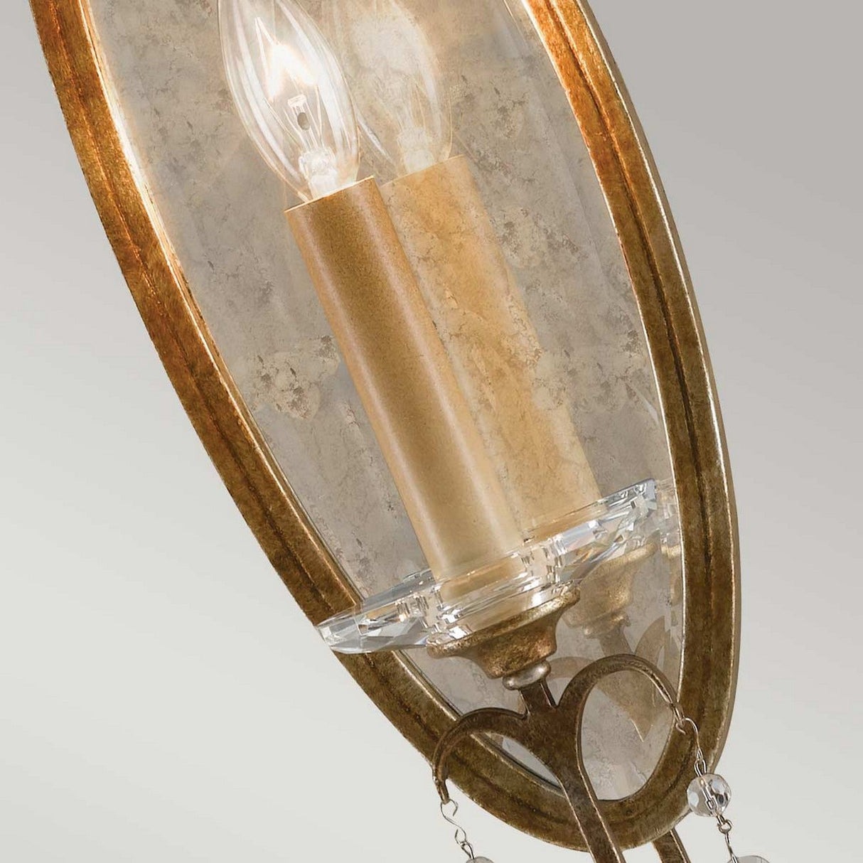 A detailed view of the Valentina 1 Light Wall Light - Oxidized Bronze showcases a candle-shaped bulb, set within a sconce featuring an oxidized bronze oval frame. It has a textured, beige stone-like backing and is mounted on a clear circular base embellished with polished glass crystals against a light gray background.