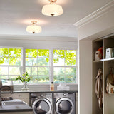 A bright laundry room features large windows with views of lush greenery outside. Inside, there is a front-loading washer and dryer with neatly folded towels on top. A countertop adorned with a vase of flowers is complemented by shelves storing baskets. The room is well-lit with the Vintner 2 Light Flush in Satin Nickel on the ceiling.