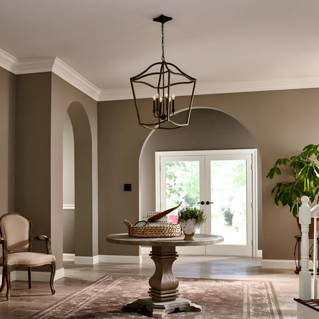 A living room with beige walls showcases a round wooden table paired with an upholstered armchair. Overhead, the Yarmouth 6 Light Chandelier in Painted Aged Brass, with its geometric design inspired by traditional lanterns, illuminates the space. A large rug spans the floor, while double doors reveal a vibrant garden view. To the right, a potted plant adds a touch of greenery.
