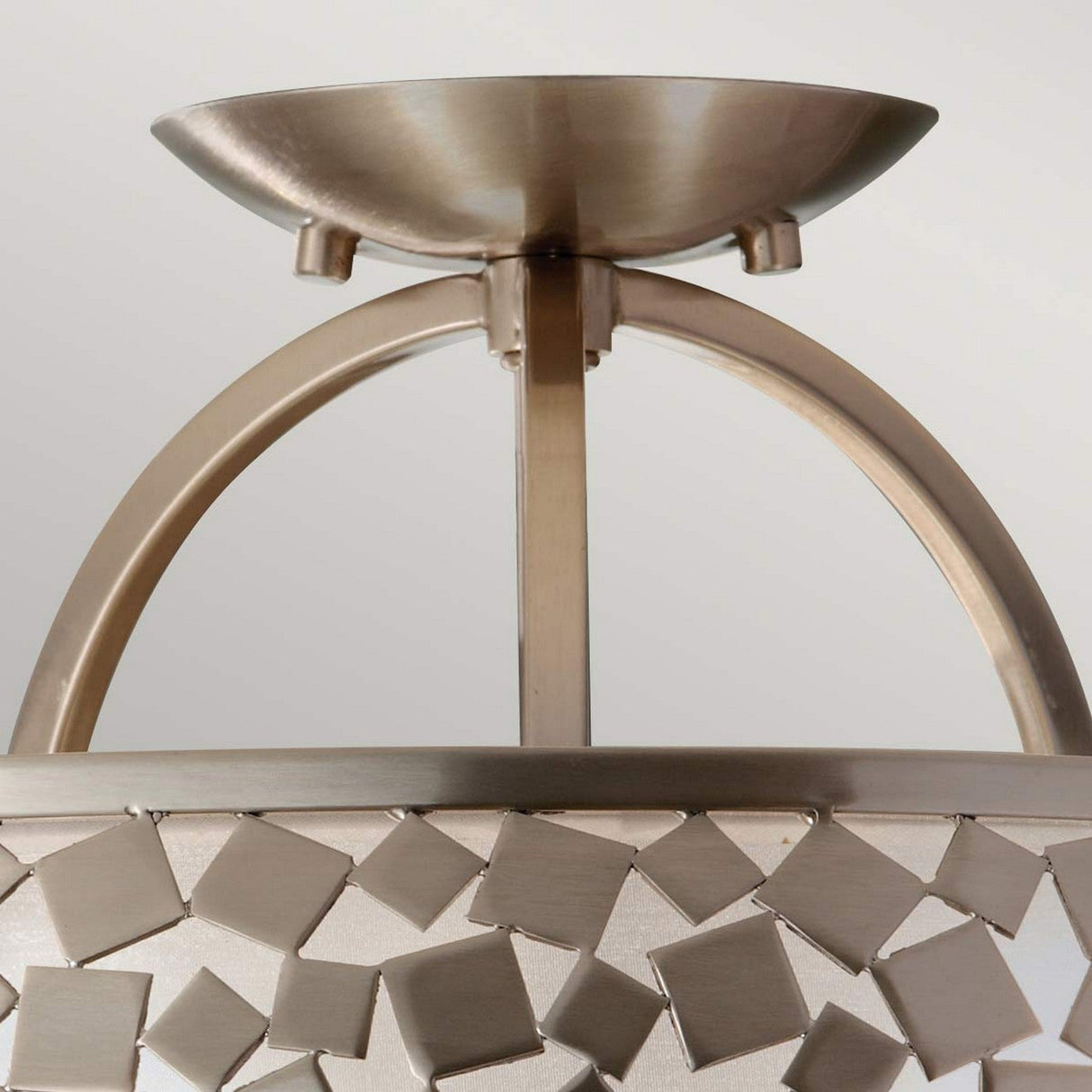 Close-up of the Zara 3 Light Semi Flush- Brushed Steel ceiling fixture highlighting its contemporary design. This modern piece is adorned with a brushed steel finish and a decorative square pattern, featuring an arched support structure that is elegantly complemented by a mosaic-like element encircling the light casing.