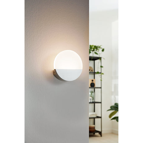 Fesessa LED Wall Light 13cm in White, a round aluminium and satin plastic disc with a half-moon lower cover, shown glowing warm white on a grey wall in a living room setting.