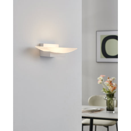 Fesessa LED Wall Light 20cm in White, a curved satin-finish shade on a slim rectangular aluminium mount, shown glowing warm white on a wall beside a dining area.