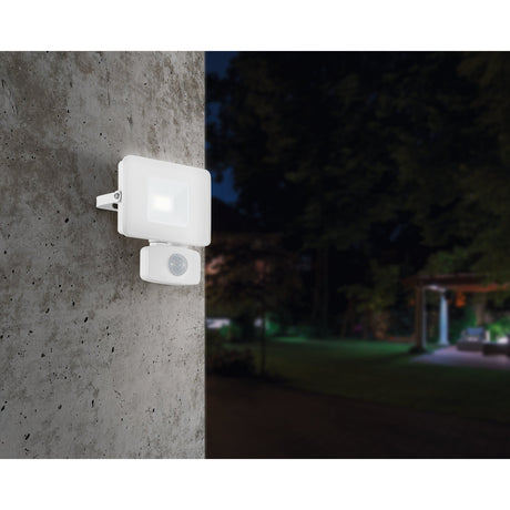 White Fesvine LED flood light with motion sensor mounted on a concrete wall, illuminating a dark garden setting at night.