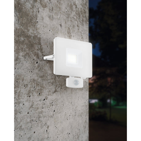 Fesvine LED flood light with motion sensor in white aluminium and glass, mounted on an outdoor concrete wall and illuminated at night.
