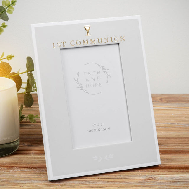 Grey freestanding First Communion photo frame with white edges, gold text and chalice icon, holding a 6 x 4in portrait photo, shown on a wooden surface with greenery and a candle.