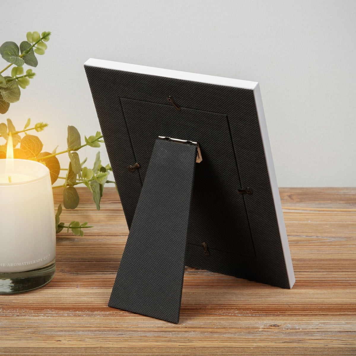 Rear view of the First Communion 6 x 4in photo frame showing the grey fabric back, hinged kickstand, and hanging tabs, set on a wooden surface beside a candle and foliage.