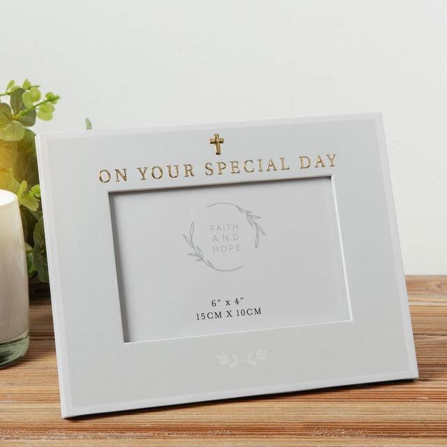Grey rectangular 6x4in photo frame with gold "On Your Special Day" text and small cross above the opening, displayed on a wooden surface beside a plant.