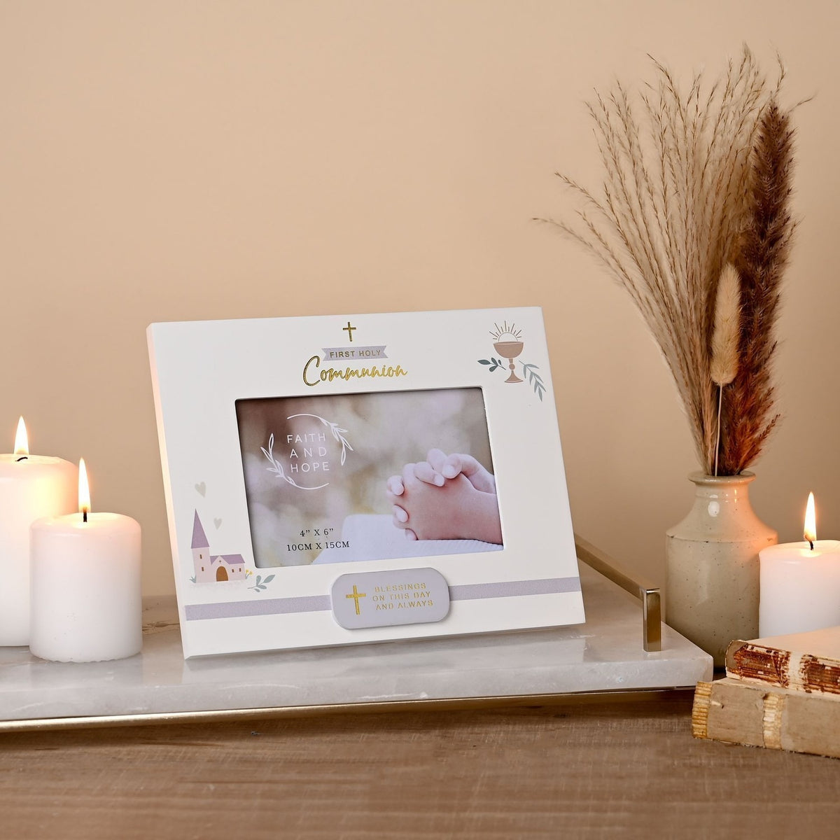 White wooden First Holy Communion photo frame with gold cross and script, chalice and church motifs, holding a photo of praying hands, displayed among candles and dried grasses.
