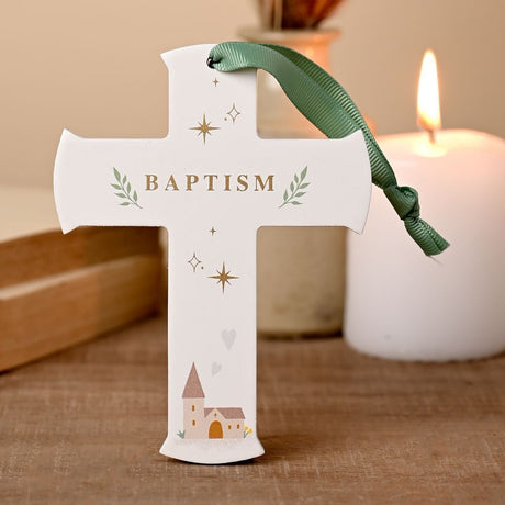White Baptism hanging cross plaque with gold "Baptism" text, stars, leaf sprigs and church illustration, sage green ribbon, shown beside a lit candle.