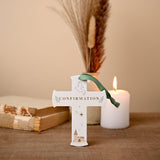 White wooden hanging cross plaque with "CONFIRMATION" in gold text, dove, stars, and church design, green ribbon, displayed by a candle and dried pampas grass.