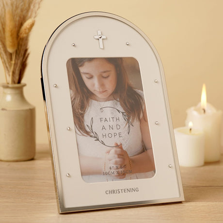 Arched Christening photo frame in silver with a cross and crystal studs, holding a girl's portrait, displayed on a table beside candles and dried flowers.