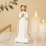 Praying Mary resin figurine in white with gold-edged veil, hands clasped and floral hair detail, on a square base beside lit candles.