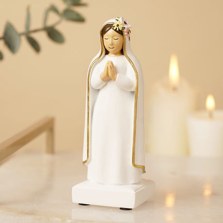 Praying Mary resin figurine in white with gold-edged veil, hands clasped and floral hair detail, on a square base beside lit candles.