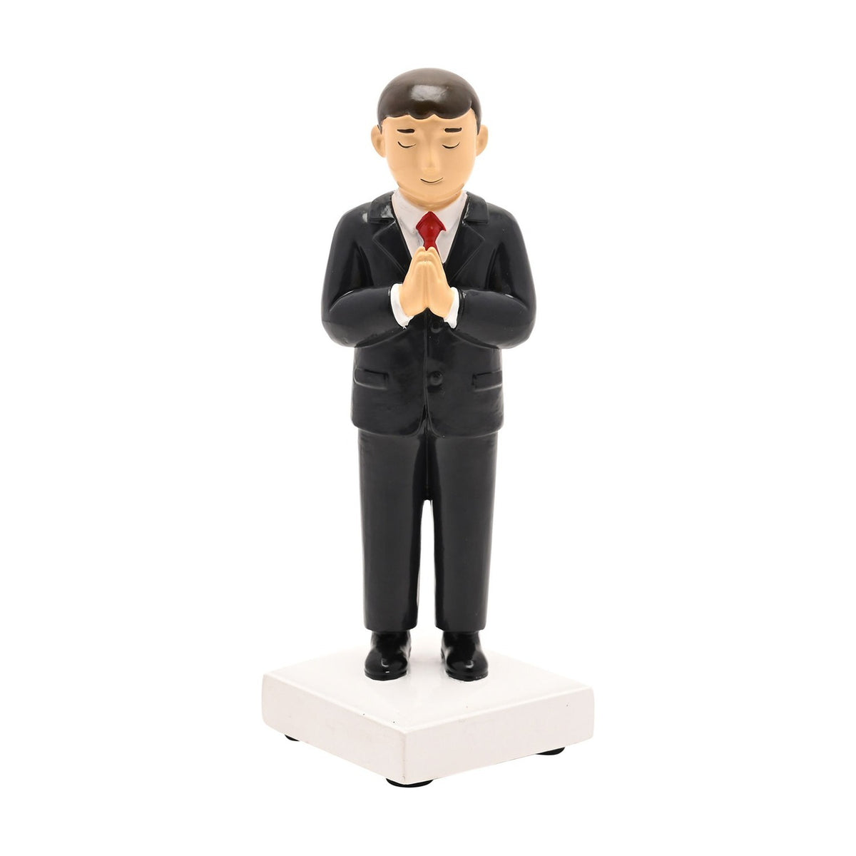 Resin Communion boy figurine with brown hair in a black suit, white shirt and red tie, hands pressed in prayer on a white square base, plain white background.