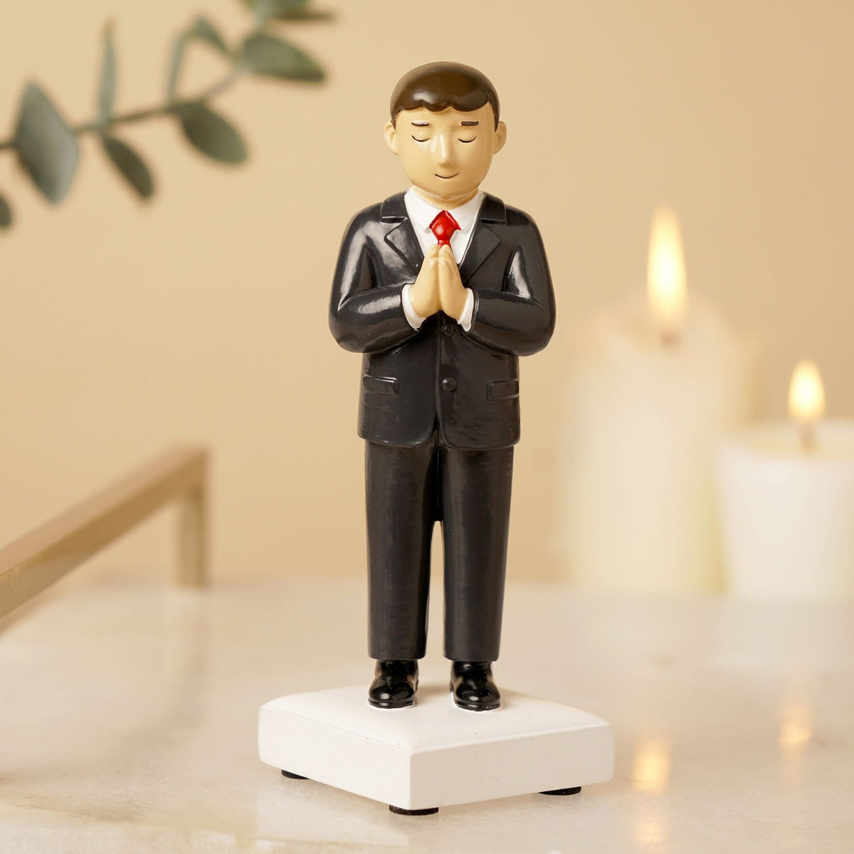 Communion Boy resin figurine in a black suit with white shirt and red tie, brown hair, hands clasped in prayer, standing on a white square base, shown on a tabletop with candles in the background.
