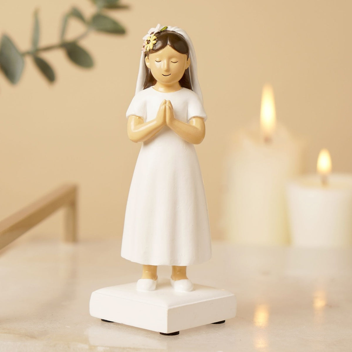 Communion Girl Figurine with dark brown hair in a white dress and veil with floral crown, hands clasped in prayer on a white base, displayed with candles in the background.