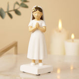 Communion Girl Figurine with dark brown hair in a white dress and veil with floral crown, hands clasped in prayer on a white base, displayed with candles in the background.