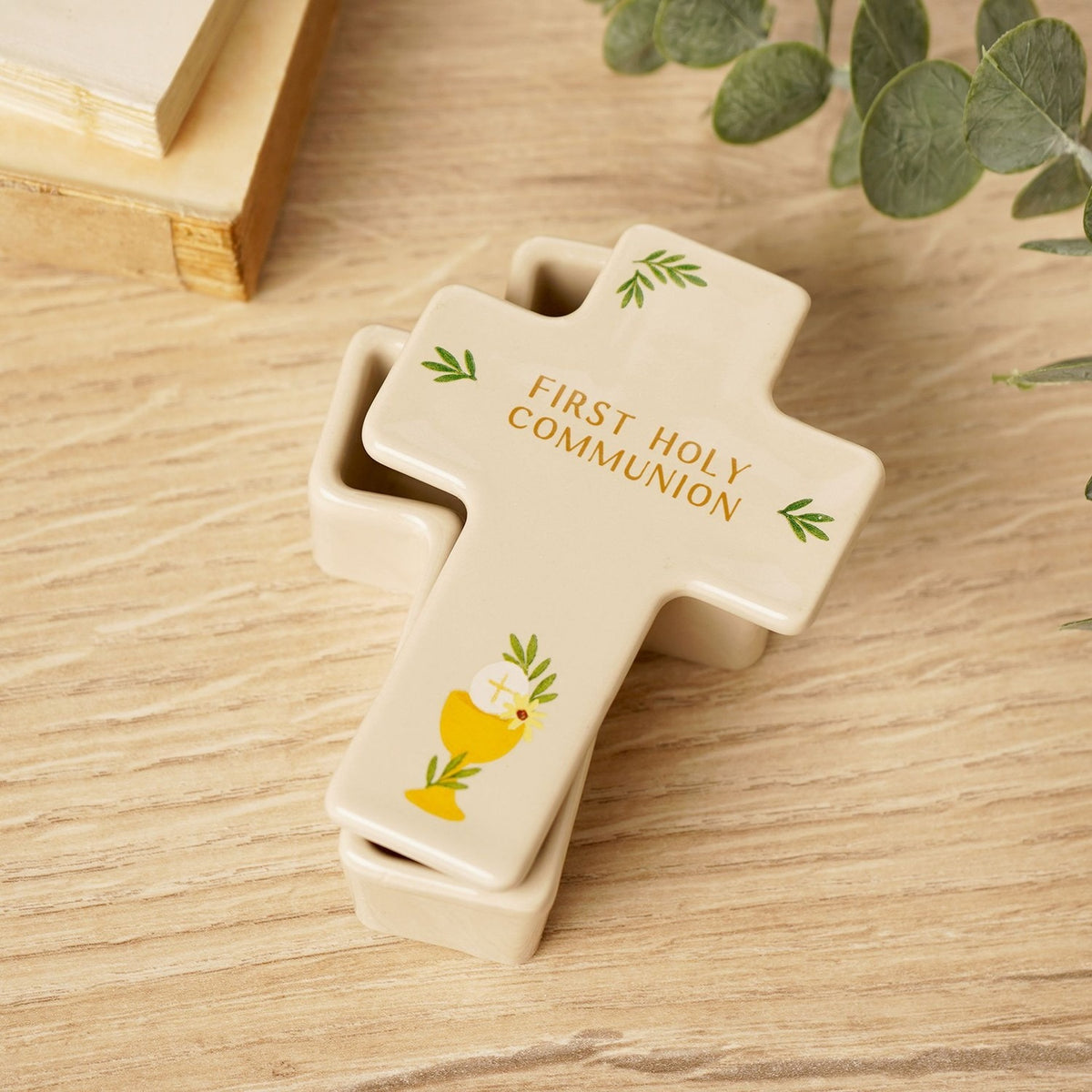 Cream ceramic cross-shaped trinket box with lid, reading "First Holy Communion" in gold with a chalice and leaf motifs, shown on a wooden surface beside greenery.