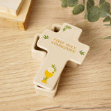 Cream ceramic cross-shaped trinket box with lid, reading "First Holy Communion" in gold with a chalice and leaf motifs, shown on a wooden surface beside greenery.