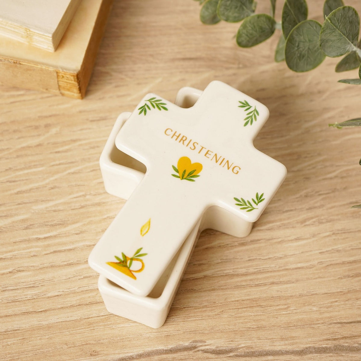 White ceramic cross-shaped trinket box with lid, "CHRISTENING" in gold lettering and green leaf, heart, and candle motifs, shown on a wooden surface with greenery.