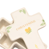 Cross-shaped white ceramic trinket box with lid, lid printed "CHRISTENING" in gold lettering above a yellow heart and green leaf motifs, shown open on a white background.