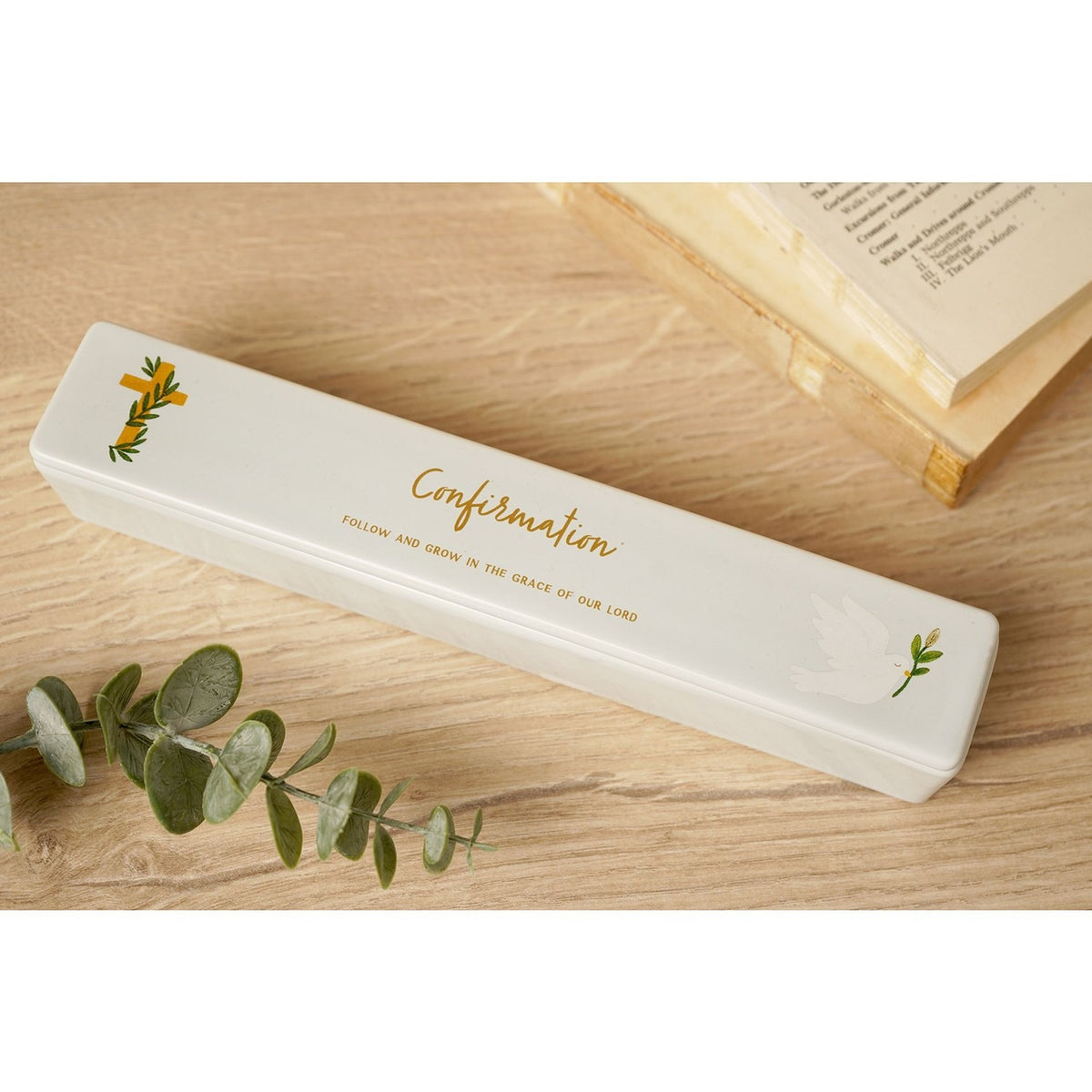 White ceramic rectangular Confirmation certificate keepsake box with gold cross and dove motifs, displayed on a wooden surface beside eucalyptus leaves and a book.